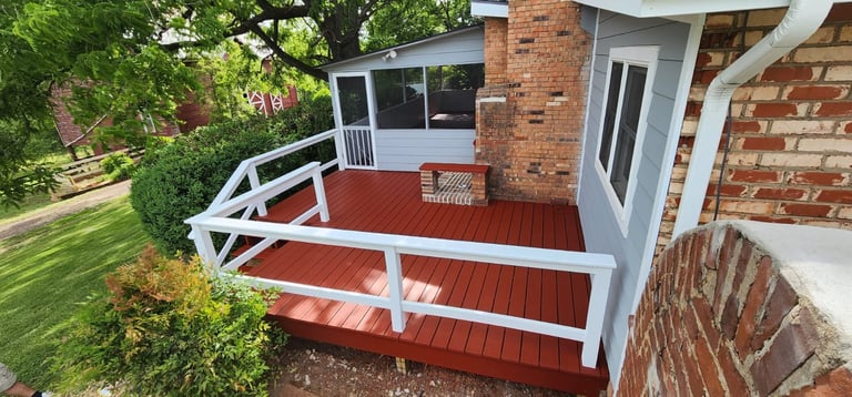 Red wooden deck with white railings attached to brick house with green lawn and landscaping visible